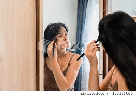 Mature 50 years old Russian woman applying contouring powder at home, enhancing her makeup while looking in the mirror, showing natural wrinkles and age beauty. Mature 50 years old Russian woman applying contouring powder at home, enhancing her makeup while looking in the mirror, showing natural wrinkles and age beauty. 133355605