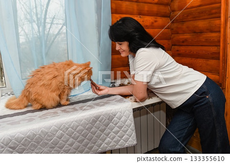 Mature 50 years old Russian woman feeds her senior 20 years old ginger cat by the window, sharing a warm and tender moment in a cozy wooden room. Mature 50 years old Russian woman feeds her senior 20 years old ginger cat by the window, sharing a warm and tender moment in a cozy wooden room. 133355610