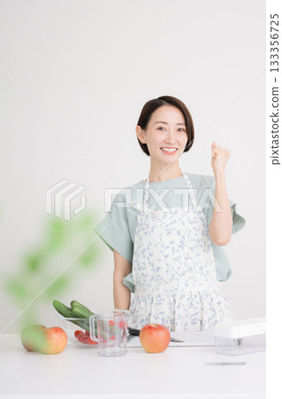 一位面帶笑容、身穿圍裙的家庭主婦在廚房準備蔬菜菜餚，並興奮地揮拳慶祝。居家飲食與健康 133356725
