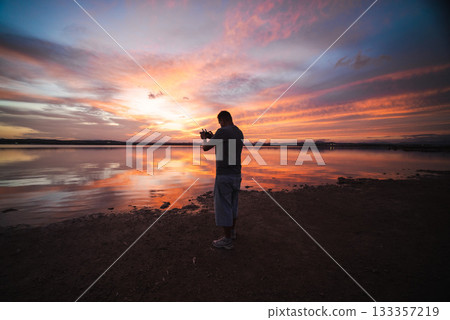 Man standing on the shore with a drone controller at sunset, beautiful reflections of orange and purple clouds on the calm lake surface. 133357219
