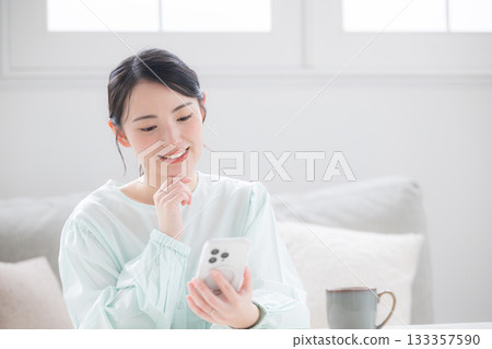 A cute Japanese woman holding a smartphone and smiling, feeling happy indoors. Image of frequently asked questions for inquiries. A cute Japanese woman holding a smartphone and smiling, feeling happy indoors. Image of frequently asked questions for inquiries. 133357590