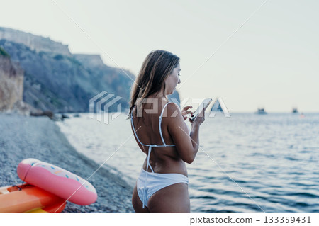 Woman smartphone beach. Young woman in white bikini using her smartphone on a pebble beach with cliffs and ocean view. Woman smartphone beach. Young woman in white bikini using her smartphone on a pebble beach with cliffs and ocean view. 133359431