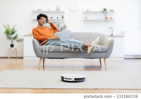 A person relaxes on a couch while a robot vacuum cleans the floor in a modern living room. 133360239