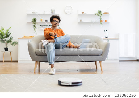 A young man watches TV while a robot vacuum cleaner cleans the floor in his modern living room. A young man watches TV while a robot vacuum cleaner cleans the floor in his modern living room. 133360250