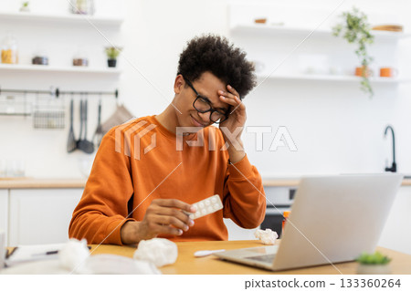 A young man experiencing a headache, holding medication, and working on a laptop in a kitchen setting. A young man experiencing a headache, holding medication, and working on a laptop in a kitchen setting. 133360264