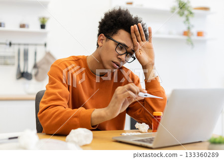 A young Black man with glasses checks his temperature while working on his laptop at home, appearing unwell. 133360289
