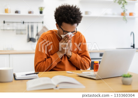 An African American man sneezes into a tissue while working from home, with a laptop and medication on the table. 133360304