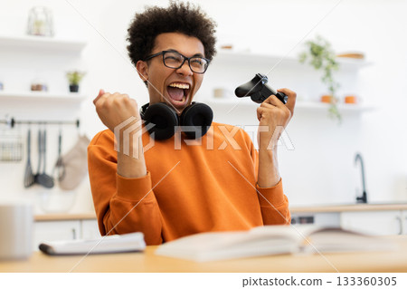 A young man with glasses celebrates a win while playing a video game, holding a controller. A young man with glasses celebrates a win while playing a video game, holding a controller. 133360305