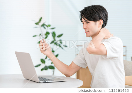 A young Asian man teleworking using a smartphone and computer in the living room of his home (troubled, pinch, trouble) 133361433