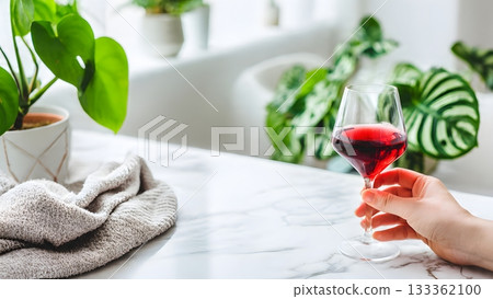 A hand gracefully holds a glass of red wine on a bright white marble kitchen countertop, surrounded by lush green indoor houseplant. AI Generated A hand gracefully holds a glass of red wine on a bright white marble kitchen countertop, surrounded by lush green indoor houseplant. AI Generated 133362100