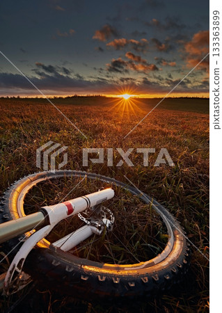 A bicycle wheel against the background of a meadow and a bright sunset sky with the sun. Travel and adventure A bicycle wheel against the background of a meadow and a bright sunset sky with the sun. Travel and adventure 133363899
