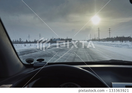 View from the windshield of a car on a snowy road with streaks of snow against the background of the sky and the sun. Dangerous driving in winter in difficult weather conditions View from the windshield of a car on a snowy road with streaks of snow against the background of the sky and the sun. Dangerous driving in winter in difficult weather conditions 133363921