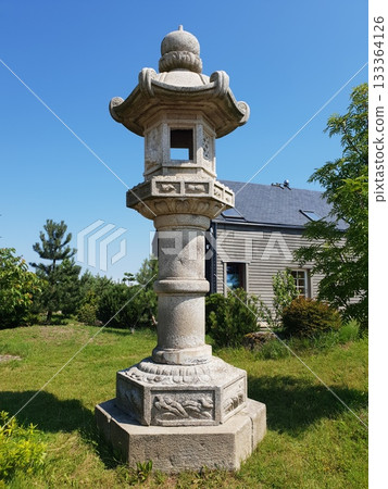 Stone lantern structure stands tall in a lush garden, surrounded by greenery and blue sky, showcasing traditional design and serene outdoor ambiance 133364126