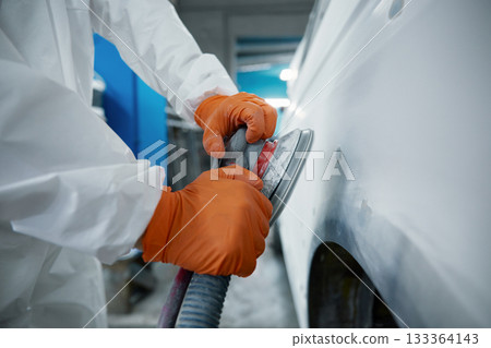 An auto body technician sands a car panel in a protective suit and orange gloves 133364143