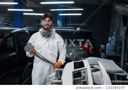 An auto body technician sands a car bumper in a paint shop, wearing safety gear 133364147