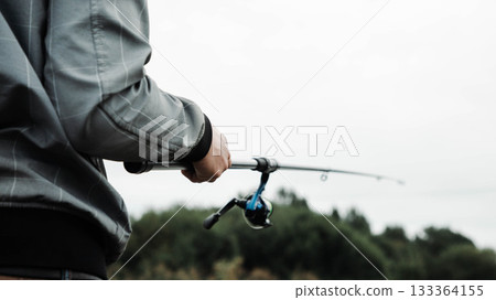 Individual holding fishing rod, wearing gray jacket, standing by water's edge, surrounded by lush greenery, enjoying peaceful outdoor activity in nature's serene environment Individual holding fishing rod, wearing gray jacket, standing by water's edge, surrounded by lush greenery, enjoying peaceful outdoor activity in nature's serene environment 133364155