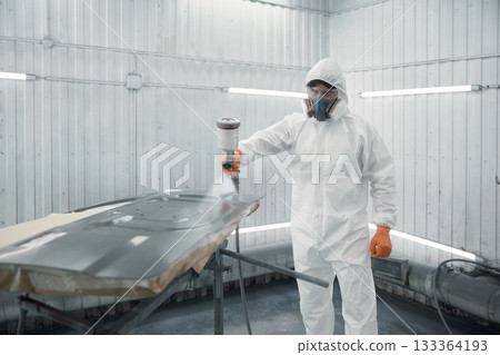 A worker dressed in a protective suit is spray-painting a car panel inside a modern paint booth 133364193