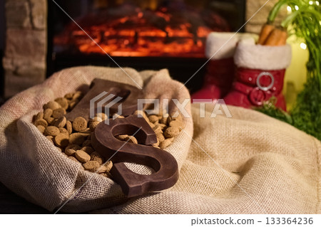 Warm Holiday Hearth. Cozy scene with chocolate letters, burlap sack, and holiday decor by the fireplace. 133364236