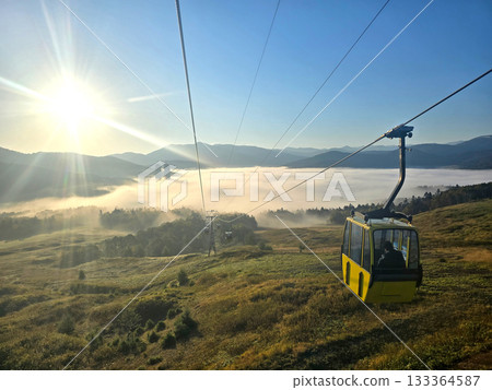 A yellow cable car ascends a mountain, offering stunning views of the valley and the morning sun A yellow cable car ascends a mountain, offering stunning views of the valley and the morning sun 133364587