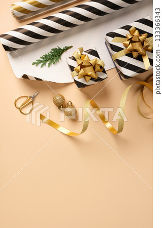 Christmas gift wrapping with golden, black and white striped wrapping paper, ribbon, scissors, and small gift boxes on beige backdrop. Copy space. Christmas gift wrapping with golden, black and white striped wrapping paper, ribbon, scissors, and small gift boxes on beige backdrop. Copy space. 133366343
