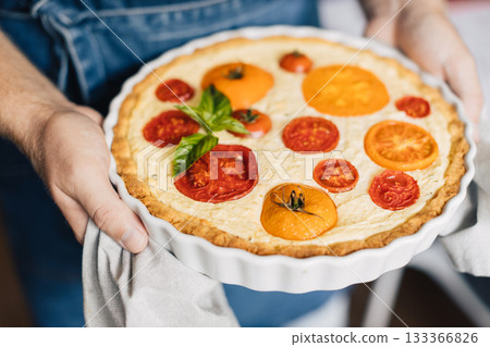 Man hands holding cheese and tomato quiche in the ceramic baking dish. Man hands holding cheese and tomato quiche in the ceramic baking dish. 133366826