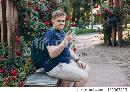 Blond European young man enjoying and listening music outside in the park. Handsome male sitting on bench and relaxing with music in Bluetooth headphones. Lifestyle, technology, people concept. Blond European young man enjoying and listening music outside in the park. Handsome male sitting on bench and relaxing with music in Bluetooth headphones. Lifestyle, technology, people concept. 133367225