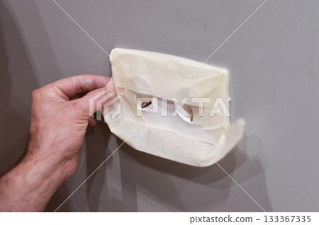 Focused on a pair of hands, this image demonstrates the practical step of masking an electrical outlet with durable tape to safeguard it from accidental paint damage during home repairs. 133367335