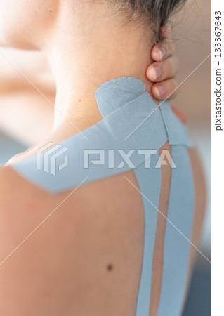 Close up view of young woman with kinesiology tape applied to her neck and shoulder holding her neck with her hand. Physical therapy and muscle recovery through modern rehabilitative care 133367643