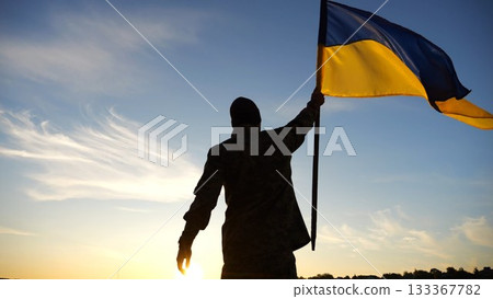 Silhouette of male military lifting blue-yellow banner in honor of the victory against russian aggression at sunset. Soldier of ukrainian army raised a waving flag of Ukraine at countryside. Slow mo 133367782