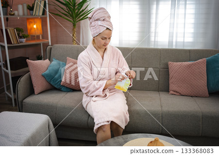 Woman wearing bathrobe opening cosmetic product sitting on sofa at home 133368083