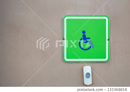 Bright green sign with symbol of person in wheelchair on the wall next to white button that, when pressed, will bring person to assist people with disabilities and physical injuries 133368658