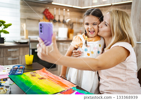 Mother kisses her daughter as they happily capture a selfie together in their kitchen Mother kisses her daughter as they happily capture a selfie together in their kitchen 133368939