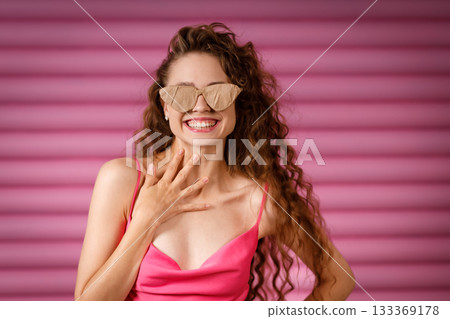 Young positive smiling woman receives a gift and holds in her hands a glasses wrapped in rustic beige craft paper on pink background. Black Friday concept. 133369178