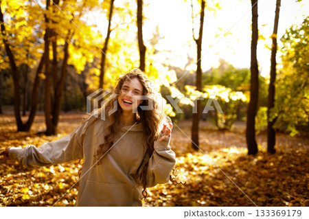 Smiling young woman plays and jumping dancing in the autumn forest with the yellow leaves at sunset 133369179