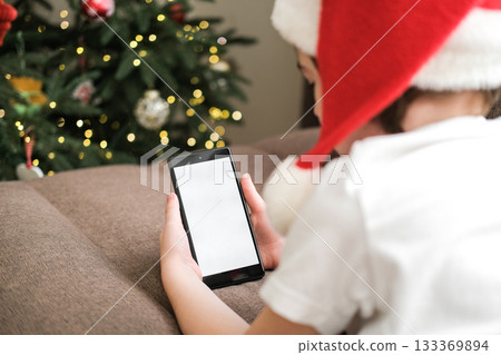 Five-year-old boy in Santa hat using a smartphone with blank white screen on sofa next to decorated Christmas tree. Christmas holidays, video calls, mobile apps, screen mockup Five-year-old boy in Santa hat using a smartphone with blank white screen on sofa next to decorated Christmas tree. Christmas holidays, video calls, mobile apps, screen mockup 133369894