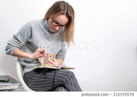 blond woman with glasses counting money and writing in a notebook, isolated on a gray 133370579
