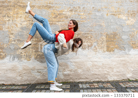 funny teenager carrying her best friend piggyback on the street at the old wall, positive emotions 133370711