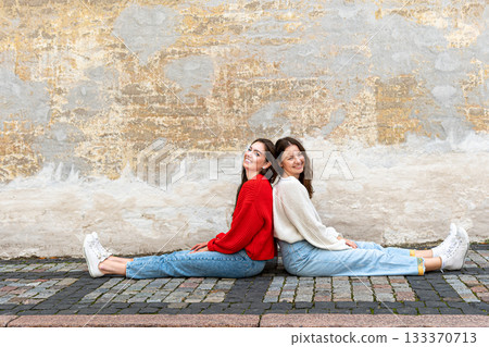 two teenagers sitting back to back on the sidewalk by an old wall 133370713