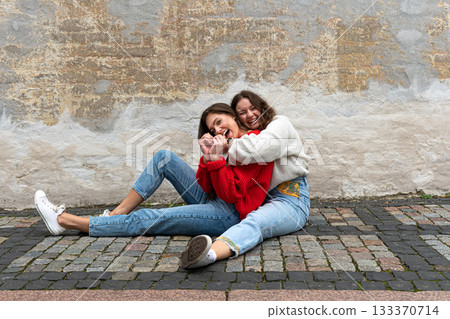 two young women hugged sitting on a paved sidewalk by an old wall 133370714