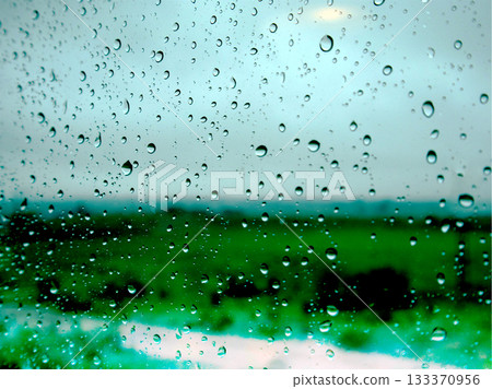 Raindrops on glass with a blurred green field and gloomy grey sky in the background.Water droplets on a window in rainy season. Raindrops on glass with a blurred green field and gloomy grey sky in the background.Water droplets on a window in rainy season. 133370956