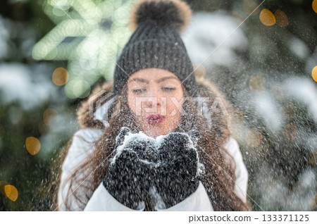 young woman blows snow from her hands while standing against the background of a Christmas tree young woman blows snow from her hands while standing against the background of a Christmas tree 133371125