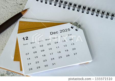 White December 2025 calendar on notepad and pen background. December month and year end concept. White December 2025 calendar on notepad and pen background. December month and year end concept. 133371510