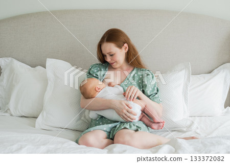 Happy family at home. Mother breastfeeding baby for health nutrition wellness. Mom nursing feeding her newborn child milk at home. Woman infant girl in white bedroom Mother hugs baby with love care 133372082