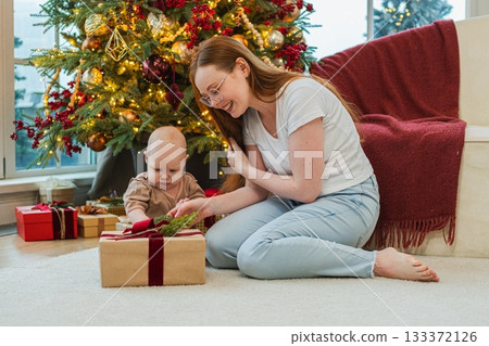 Merry Christmas Xmas. Happy family mother newborn baby child unpacking gift box near Christmas tree at home. Mom girl kid celebrating first Christmas enjoying winter holidays together. Family moments Merry Christmas Xmas. Happy family mother newborn baby child unpacking gift box near Christmas tree at home. Mom girl kid celebrating first Christmas enjoying winter holidays together. Family moments 133372126