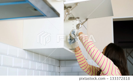 A young woman focuses on screwing a screw into a cabinet during kitchen assembly wearing gloves and working carefully beneath the cabinet in a bright home environment, renovation concept  133372818