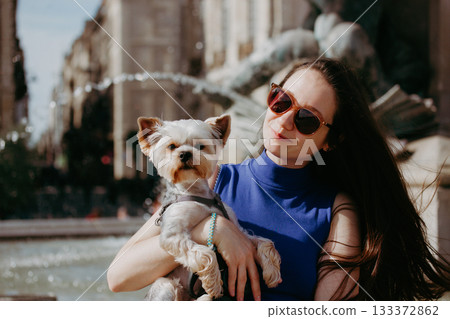 Young brunette woman holding a small dog Yorkshire terrier in front of a fountain. A girl with sunglasses holds a doggy in her arms, posing on a city street on a sunny day. A trip with a lapdog, pet. 133372862