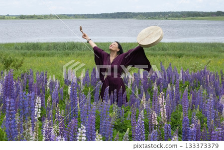 Shaman woman drumming among blooming flowers in summer 133373379