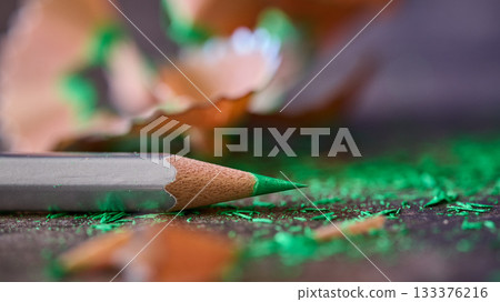 A green pencil rests next to wood shavings on a surface. 133376216