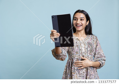 Confident upbeat girl sharing a story with best friend over web video call, standing against blue background with smartphone. Happy woman talking with mate online on teleconference. 133377577