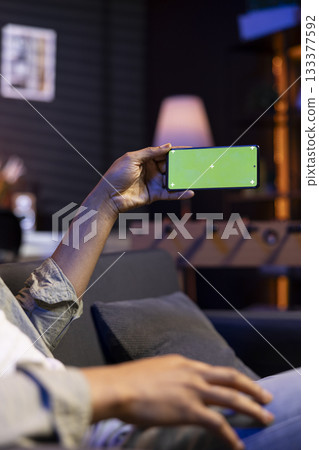 African american young adult laying on the sofa with mockup screen, feeling happy and relaxed with flexible work schedule at home. Male freelancer uses his phone relaxing in the living room. African american young adult laying on the sofa with mockup screen, feeling happy and relaxed with flexible work schedule at home. Male freelancer uses his phone relaxing in the living room. 133377592